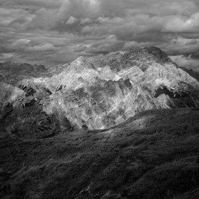 A dramatic black and white photograph of a rugged mountain range under a cloud-filled sky. The peaks are sharply defined with contrasting light and shadow, adding depth and texture to the scene. The clouds above add a sense of impending weather and movement.