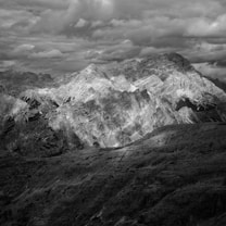 A dramatic black and white photograph of a rugged mountain range under a cloud-filled sky. The peaks are sharply defined with contrasting light and shadow, adding depth and texture to the scene. The clouds above add a sense of impending weather and movement.