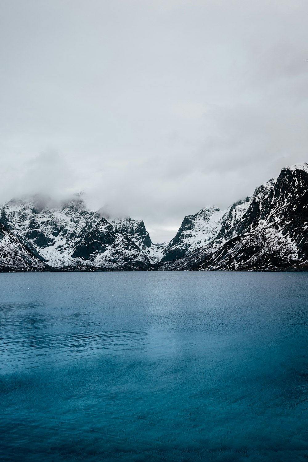 Plan d’eau bleu calme et montagne enneigée sous les nuages gris pendant la journée