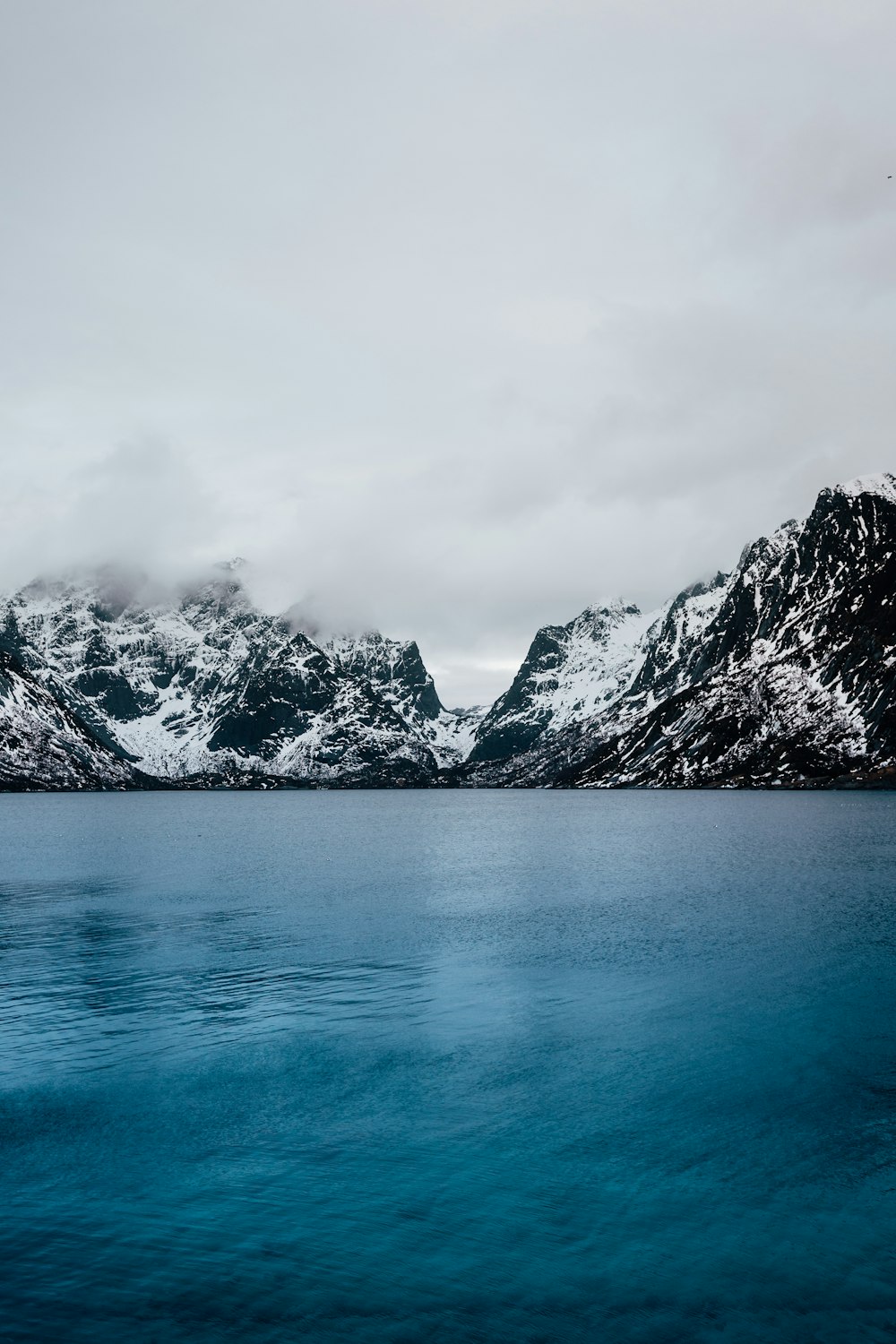 blaues, ruhiges Gewässer und schneebedeckter Berg unter grauen Wolken am Tag