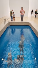 A shallow pool with crystal-clear blue water reflects the figures of people standing at the edge. The pool is indoors, surrounded by a minimalistic, white-walled environment with wooden flooring. Some people are looking down into the pool while others are engaged in conversation.