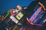 A vibrant urban street scene with illuminated neon signs, including a large guitar and various text emphasizing music themes. The atmosphere suggests nightlife with bright, colorful lights against a darkening sky. The signs promote a record shop and highlight live music culture.
