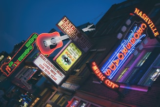 A vibrant urban street scene with illuminated neon signs, including a large guitar and various text emphasizing music themes. The atmosphere suggests nightlife with bright, colorful lights against a darkening sky. The signs promote a record shop and highlight live music culture.