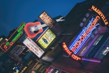 A vibrant urban street scene with illuminated neon signs, including a large guitar and various text emphasizing music themes. The atmosphere suggests nightlife with bright, colorful lights against a darkening sky. The signs promote a record shop and highlight live music culture.