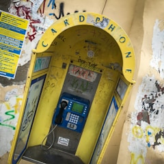 A weathered public phone booth with a prominently displayed yellow arch labeled 'CARD PHONE'. The booth is covered with various graffiti and stickers. It houses a blue payphone with a traditional handset and keypad. Surrounding walls are adorned with more graffiti and a blue informational poster.