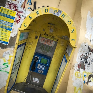 A weathered public phone booth with a prominently displayed yellow arch labeled 'CARD PHONE'. The booth is covered with various graffiti and stickers. It houses a blue payphone with a traditional handset and keypad. Surrounding walls are adorned with more graffiti and a blue informational poster.