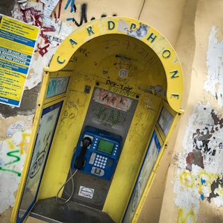 A weathered public phone booth with a prominently displayed yellow arch labeled 'CARD PHONE'. The booth is covered with various graffiti and stickers. It houses a blue payphone with a traditional handset and keypad. Surrounding walls are adorned with more graffiti and a blue informational poster.