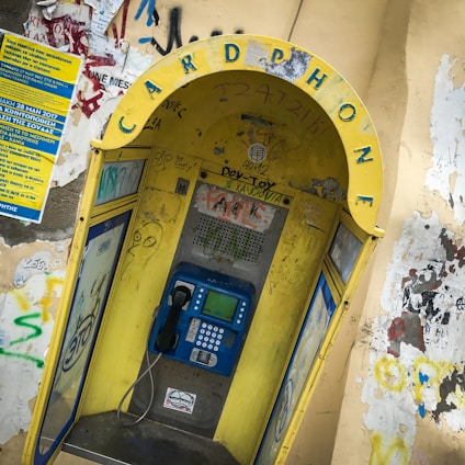 A weathered public phone booth with a prominently displayed yellow arch labeled 'CARD PHONE'. The booth is covered with various graffiti and stickers. It houses a blue payphone with a traditional handset and keypad. Surrounding walls are adorned with more graffiti and a blue informational poster.
