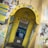 A weathered public phone booth with a prominently displayed yellow arch labeled 'CARD PHONE'. The booth is covered with various graffiti and stickers. It houses a blue payphone with a traditional handset and keypad. Surrounding walls are adorned with more graffiti and a blue informational poster.