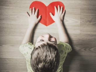 boy in green shirt holding red paper heart cutout on brown table