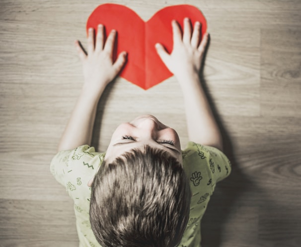 boy in green shirt holding red paper heart cutout on brown table