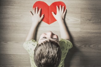 boy in green shirt holding red paper heart cutout on brown table