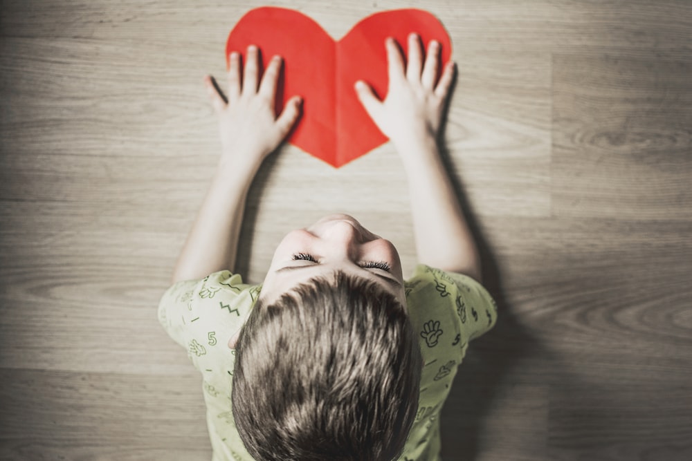 Boy in a green shirt holding a red paper heart cutout on a brown table.