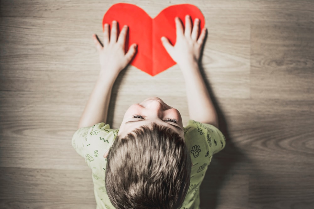 boy in green shirt holding red paper heart cutout on brown table