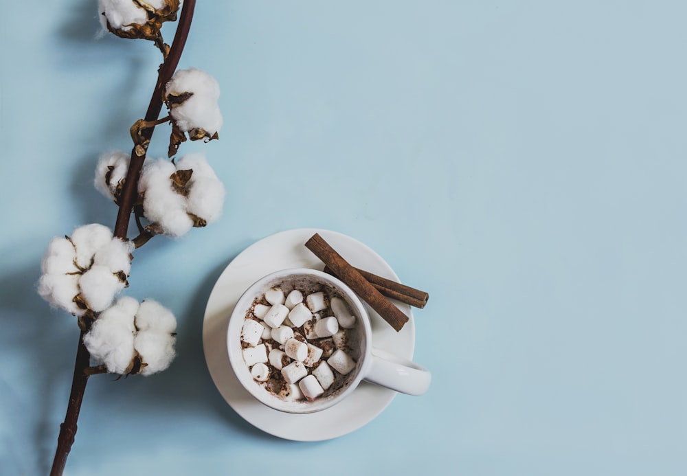 white ceramic mug filled with marshmallows beside cinnamon sticks placed on round white ceramic saucer near white cuttons