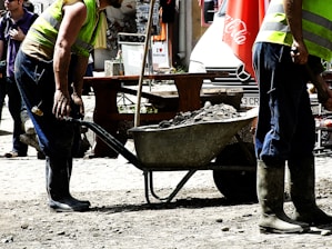 Construction workers wearing high-visibility vests and boots are using a wheelbarrow filled with rocks or debris. The scene suggests an outdoor setting with a mix of construction and urban elements. In the background, there is a street scene with a parked vehicle and various objects like a Coca-Cola umbrella.