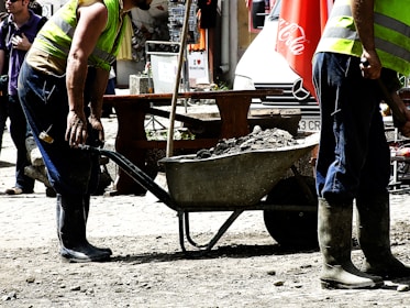 Construction workers wearing high-visibility vests and boots are using a wheelbarrow filled with rocks or debris. The scene suggests an outdoor setting with a mix of construction and urban elements. In the background, there is a street scene with a parked vehicle and various objects like a Coca-Cola umbrella.