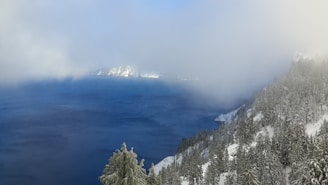 A large, deep blue lake is surrounded by snow-covered trees and hills. Fog partially obscures the scene, creating a mystical and calm atmosphere. The landscape features a dense forest blanketed in snow, with hints of distant mountains.