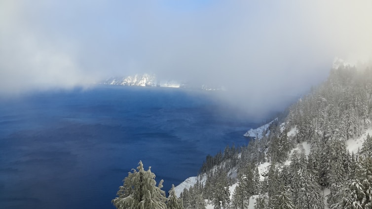 A large, deep blue lake is surrounded by snow-covered trees and hills. Fog partially obscures the scene, creating a mystical and calm atmosphere. The landscape features a dense forest blanketed in snow, with hints of distant mountains.