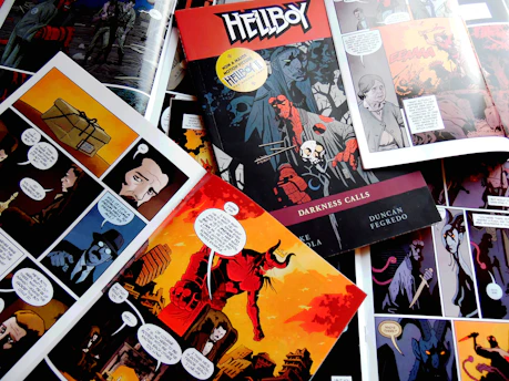 Various comic book pages are spread out featuring dark and colorful graphic art. One prominent cover titled 'Hellboy: Darkness Calls' is visible, showcasing a red figure with a stone hand holding a spear, surrounded by a skull and mysterious characters. Each page includes dialogue bubbles and depicts scenes with shadowy figures and vibrant landscapes.