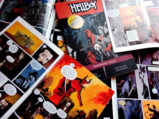 Various comic book pages are spread out featuring dark and colorful graphic art. One prominent cover titled 'Hellboy: Darkness Calls' is visible, showcasing a red figure with a stone hand holding a spear, surrounded by a skull and mysterious characters. Each page includes dialogue bubbles and depicts scenes with shadowy figures and vibrant landscapes.