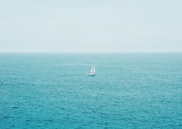 A lone sailboat cutting through deep blue ocean waves under a vast, cloudy sky.