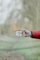 A hand wearing a red jacket sleeve holds a white enamel mug with the words 'Adventure Begins' printed on it. Liquids splash out from the mug, captured mid-air, creating an energetic and dynamic scene. The background is blurred, suggesting an outdoor setting.