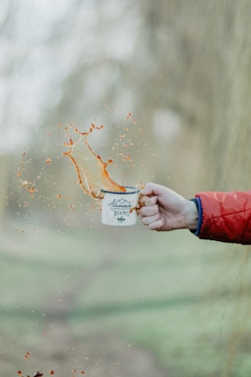 A hand wearing a red jacket sleeve holds a white enamel mug with the words 'Adventure Begins' printed on it. Liquids splash out from the mug, captured mid-air, creating an energetic and dynamic scene. The background is blurred, suggesting an outdoor setting.