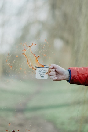 A hand wearing a red jacket sleeve holds a white enamel mug with the words 'Adventure Begins' printed on it. Liquids splash out from the mug, captured mid-air, creating an energetic and dynamic scene. The background is blurred, suggesting an outdoor setting.