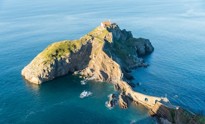 A stunning coastal landscape featuring an islet connected to the mainland by a winding stone bridge. At the highest point of the rocky outcrop sits a small structure with terracotta roofing. The surrounding sea is calm and stretches to the horizon, offering a serene and scenic view.