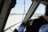 The image depicts the interior of an airplane cockpit with a pilot visible on the right. The view through the cockpit window shows a runway and several planes in the distance under a clear blue sky. Various cockpit controls and instruments are visible, along with a piece of paper with printed information attached near the window.
