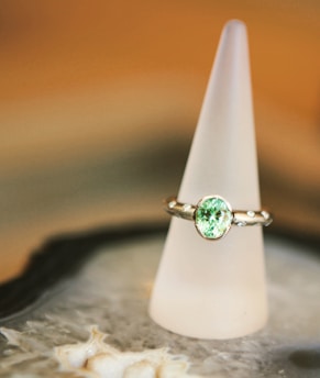 A silver ring with a prominent light green gemstone is elegantly displayed on a frosted conical stand. The background features soft, warm tones, creating a luxurious and refined atmosphere. The surface beneath the stand appears to be intricate and textured.