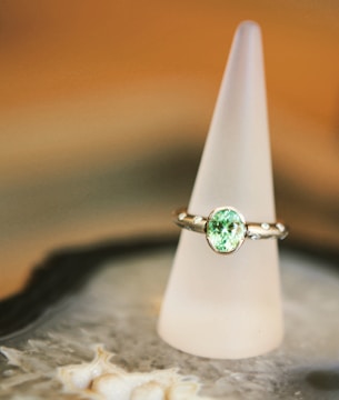A silver ring with a prominent light green gemstone is elegantly displayed on a frosted conical stand. The background features soft, warm tones, creating a luxurious and refined atmosphere. The surface beneath the stand appears to be intricate and textured.