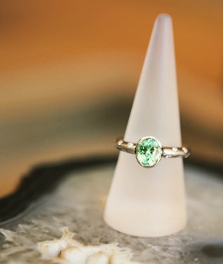 A silver ring with a prominent light green gemstone is elegantly displayed on a frosted conical stand. The background features soft, warm tones, creating a luxurious and refined atmosphere. The surface beneath the stand appears to be intricate and textured.