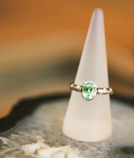 A silver ring with a prominent light green gemstone is elegantly displayed on a frosted conical stand. The background features soft, warm tones, creating a luxurious and refined atmosphere. The surface beneath the stand appears to be intricate and textured.
