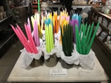 A display of colorful taper candles in various shades of pink, green, orange, yellow, and purple are arranged in containers lined with white tissue paper on a marble or stone countertop. The background features shelves with various items and a person standing at a counter.