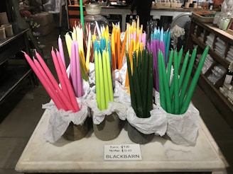 A display of colorful taper candles in various shades of pink, green, orange, yellow, and purple are arranged in containers lined with white tissue paper on a marble or stone countertop. The background features shelves with various items and a person standing at a counter.