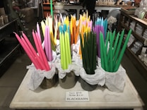A display of colorful taper candles in various shades of pink, green, orange, yellow, and purple are arranged in containers lined with white tissue paper on a marble or stone countertop. The background features shelves with various items and a person standing at a counter.
