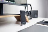 A modern workspace is depicted with a sleek black smartphone leaning against the base of a stylish wooden monitor stand, which supports a large screen displaying graphics. In the background, black headphones and a square speaker are visible, adding to the tech-inspired setting. The desk appears minimalist, with a mouse pad and computer mouse in view, indicating an organized and neat environment.