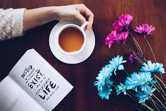 Alex Muck sitting by a towering bookshelf with a warm smile, a cup of tea beside her, absorbing a quiet moment of inspiration.