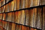 A close-up view of a wooden shingle wall, showing overlapping wooden tiles with varied shades of brown, orange, and grey. The texture appears weathered and aged, indicating exposure to natural elements.