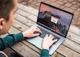 A person is using a laptop on a wooden outdoor table. The laptop screen displays an operating system desktop with several icons and a notification. The person is wearing a green sweater and their hands are on the keyboard.