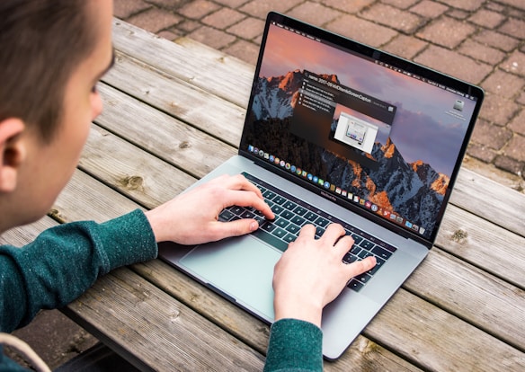 A person is using a laptop on a wooden outdoor table. The laptop screen displays an operating system desktop with several icons and a notification. The person is wearing a green sweater and their hands are on the keyboard.
