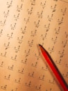 A math worksheet filled with various multiplication and division problems is accompanied by a red pencil resting on the paper. The writing appears to be handwritten, with some numbers scribbled or crossed out as part of corrections or workings.