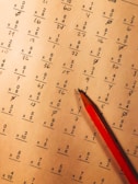 A math worksheet filled with various multiplication and division problems is accompanied by a red pencil resting on the paper. The writing appears to be handwritten, with some numbers scribbled or crossed out as part of corrections or workings.