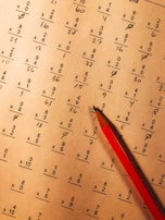A math worksheet filled with various multiplication and division problems is accompanied by a red pencil resting on the paper. The writing appears to be handwritten, with some numbers scribbled or crossed out as part of corrections or workings.