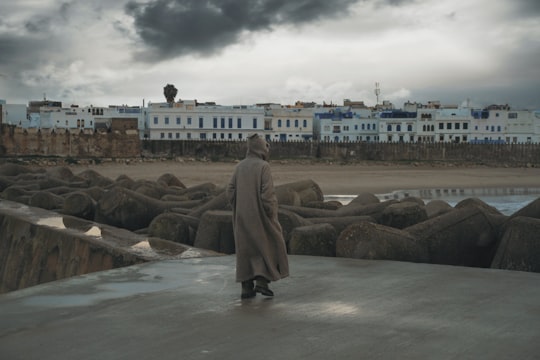 woman in brown coat standing on gray rock near body of water during daytime in Asilah Morocco
