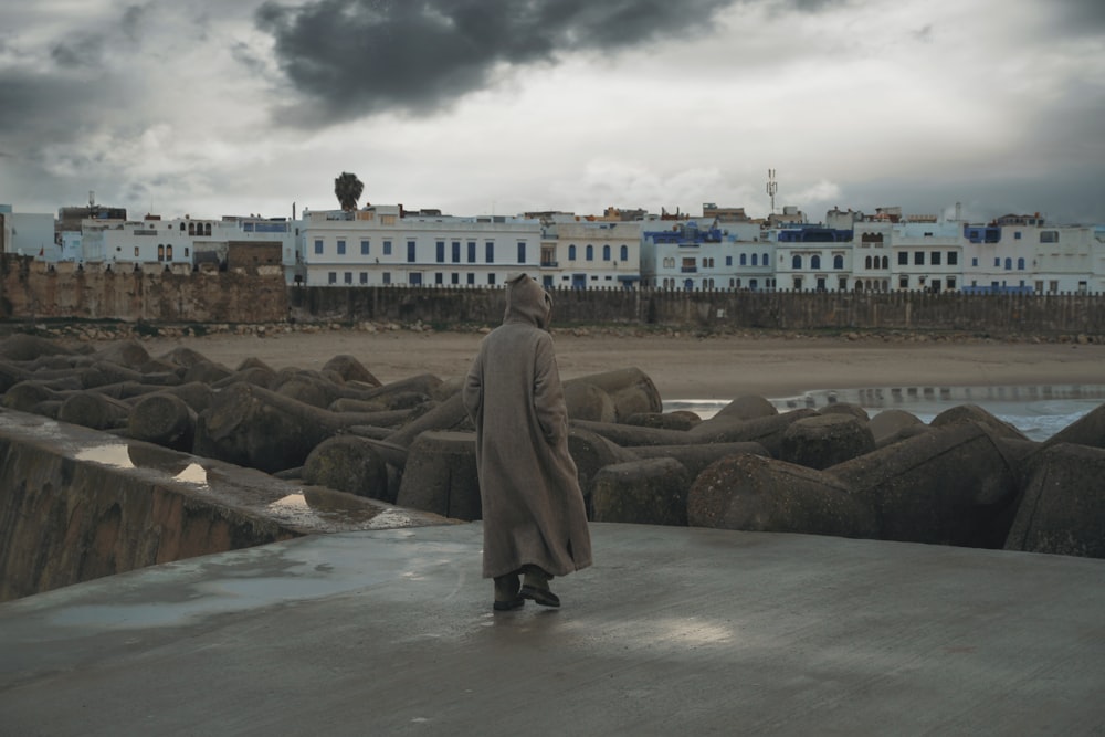 woman in brown coat standing on gray rock near body of water during daytime