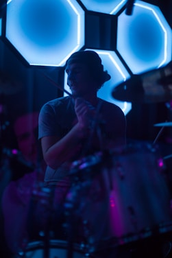 A drummer is positioned in front of a set of illuminated, hexagonal LED lights that cast a vibrant blue glow. The scene is dark, with subtle hints of purple and pink lighting adding depth. The drummer wears a backwards cap and is partially silhouetted by the bright lights behind.