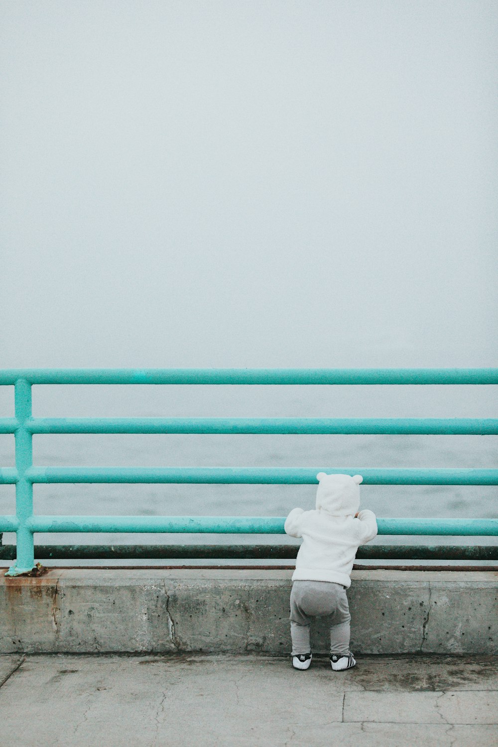 toddler standing while resting arm on rail facing body of water